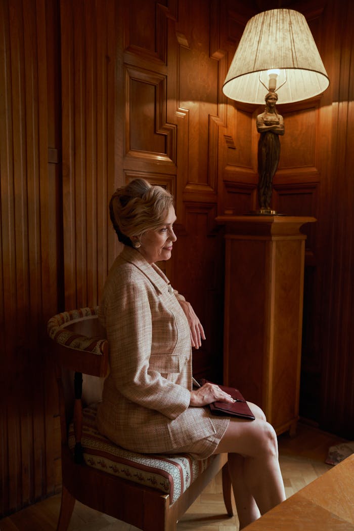 Senior woman in elegant attire seated in a classic wooden room, radiating grace.