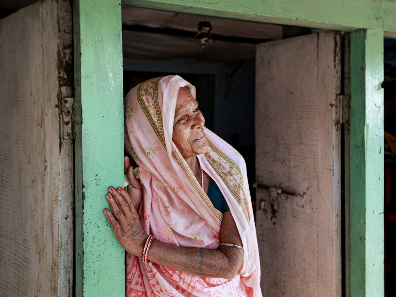 who-we-are A senior woman in a traditional sari smiles while leaning in a doorway.