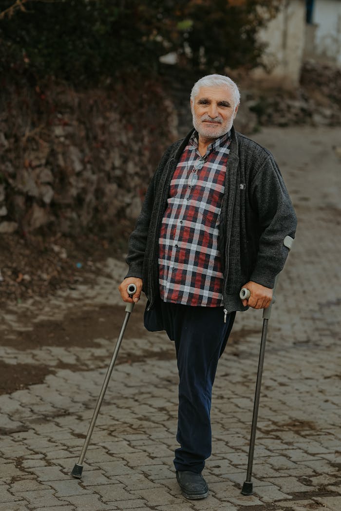 Senior man with gray hair standing on a cobblestone path using crutches outdoors.