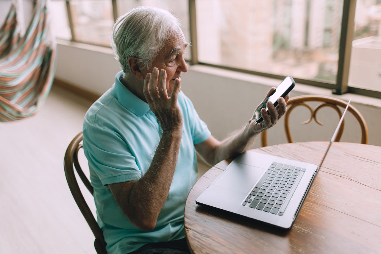 Elderly man using smartphone and laptop indoors, looking puzzled, showcasing technology and leisure.