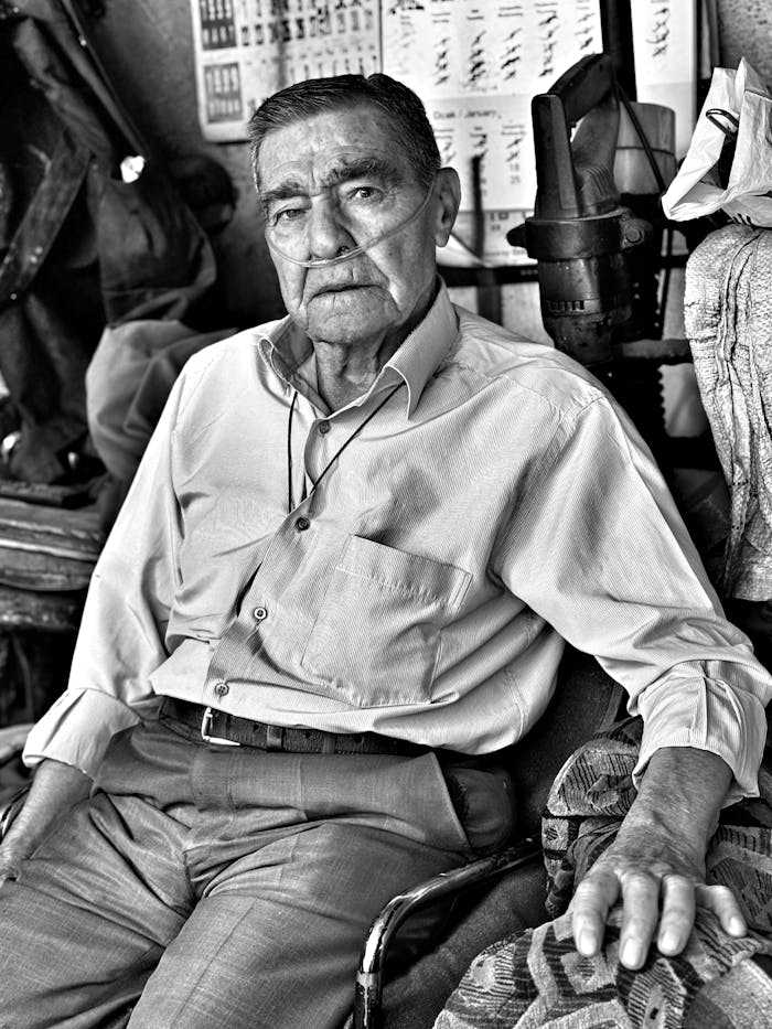 Black and white portrait of a senior man with an oxygen tube sitting in his workshop.